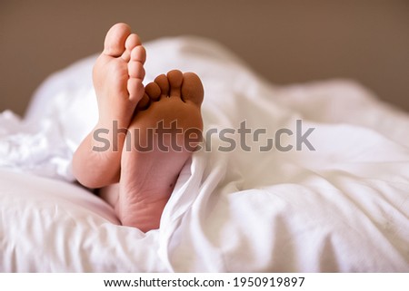 woman's leg looks out from under the covers on a white bed. Legs of a young woman in bed under a white blanket.