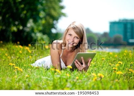 Portrait of young beautiful smiling woman with tablet pc, outdoors. Beautiful young girl lying on the grass in the field. Smiling trendy stylish woman on nature
