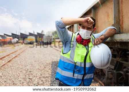 A Railway engineer or Rail transport technician wearing a green safety vest is standing to rest or working outdoors beside a freight train on a hot and sunny day. Royalty-Free Stock Photo #1950992062