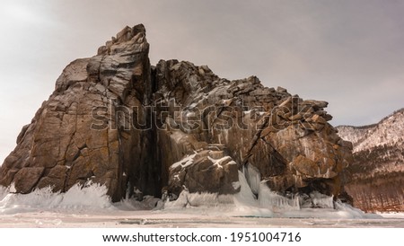 A picturesque rock without vegetation, with steep slopes and many cracks rises on a frozen lake. At the base of the cliff there are ice splashes, icicles. Sepia shades. Baikal Royalty-Free Stock Photo #1951004716