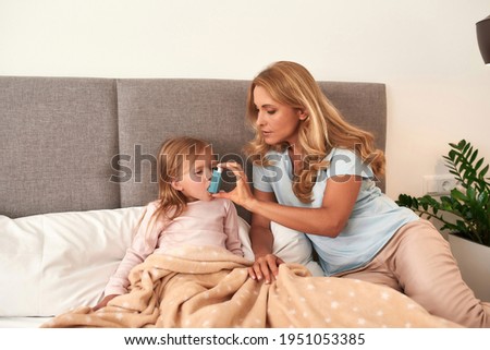 Portrait of a mother curing her daughters's asthma attack, both in bed with a plant on the background Royalty-Free Stock Photo #1951053385