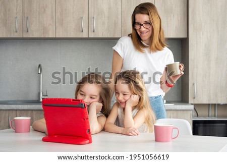 Two sisters and their mom or nanny watching a fun educational video on the tablet on the modern kitchen. Online Education concept.