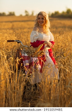 Young beautiful girl with a cute smile in a white dress with a red ribbon and a vintage bike with a basket of bread and milk in a wheat field
