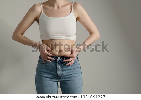 Woman in jeans and white shirt squeezing her belly fat. Woman`s hips closeup raw studio shot in grey background. Dieting and fat loss concept. Royalty-Free Stock Photo #1951061227