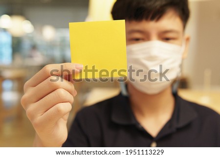 Portrait of an Asian americans young teenager boy with face mask hold a blank yellow paper note look at camera. Background, mock up, Backdrop, Medical health care, Awareness, Campaign, Stop asian hate