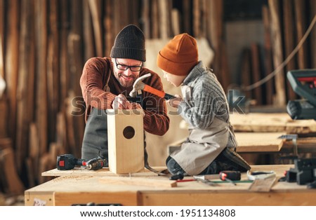 Focused little boy using hammer while making wooden bird house with father in professional joinery workshop Royalty-Free Stock Photo #1951134808