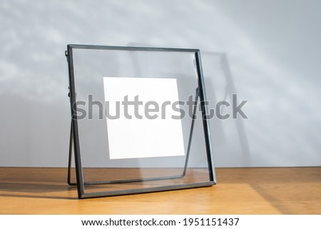 Mockup of black metallic square frame standing on wooden desk against a white wall under sunbeams. There are transparent glass installed instead of passepartout