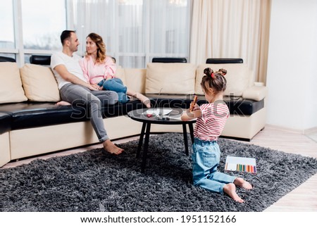 Couple sitting on sofa and little daughter drawing, daughter have a fun on warm carpet floor with colorful pencils. leisure activity at modern comfortable home concept