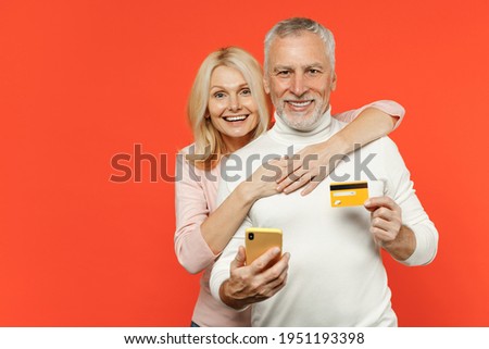 Cheerful couple two friends elderly gray-haired man blonde woman in white pink clothes using mobile cell phone hold credit bank card hugging isolated on bright orange color background studio portrait