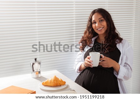 Joyful pregnant young beautiful woman looks at camera during her morning breakfast with coffee and croissants. Concept of good morning and waiting for meeting with a baby. Copyspace