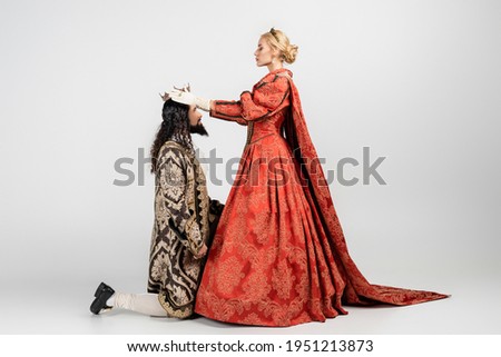full length of blonde queen wearing crown on hispanic king in medieval clothing standing on knee on white