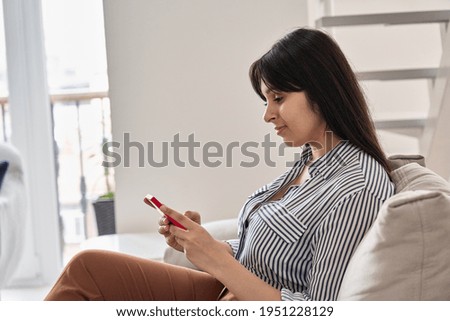Happy young indian woman using smart phone sitting on couch at home. Smiling lady holding cellphone doing online shopping, ordering delivery in smartphone application, subscribing new social media.