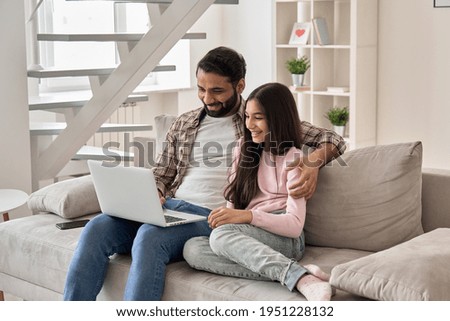 Happy indian family father with teenage child daughter having fun using laptop computer at home watching tv movie, browsing internet, e learning, having video zoom call sit on sofa in living room