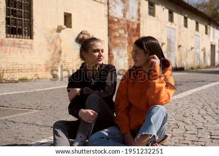 Two teenage skater girls hang out in the neighborhood. They are chatting and smiling.