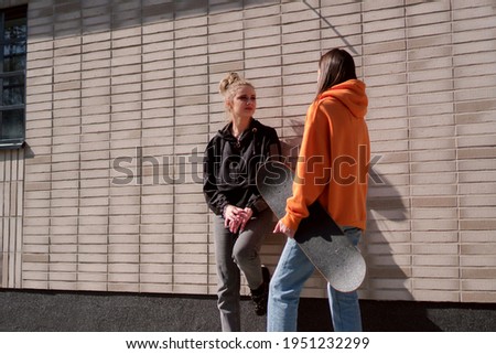 Two teenage skater girls hang out in the neighborhood. They are leaning against the wall, chatting and smiling.