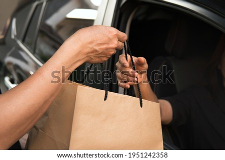 Woman’s driver driving thru hand pickup environmentally friendly craft brown paper bag out of a black car open window from seller meeting social distancing requirements and supporting small business Royalty-Free Stock Photo #1951242358