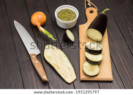 Sliced eggplant on cutting board. Mung bean groats in bowl. Knife and bow on table. Dark wooden background. Top view
