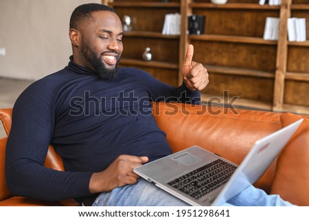 Smiling African-American man talking online via video call on the laptop in a relaxed atmosphere, a black guy lying on the couch and showing thumbs up at webcam in sign of good connection