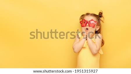 Little child girl in striped swimsuit and red funny summer sunglasses surprised expression looks at camera posing on yellow background, studio portrait.Advertising of children's products and sale