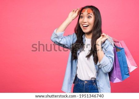 Photo of beautiful emotional surprised young cute asian woman posing isolated on pink wall background in denim clothes, holding shopping bags. Concept for discounts, sale, black friday. Copy space.