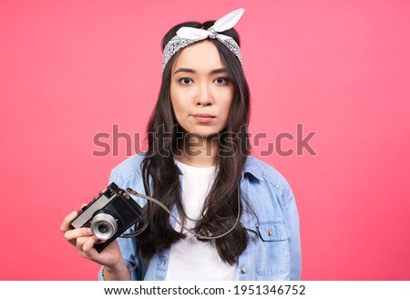 Image of a young sad disappointed young Asian girl, in casual stylish denim clothes, standing isolated on a pink background, taking pictures with a retro vintage camera.