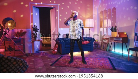 Relaxed young African American male in living room listening to music in headphones and dancing, moving body rhythmically in neon disco light in vintage apartment. Retro style, leisure concept Royalty-Free Stock Photo #1951354168