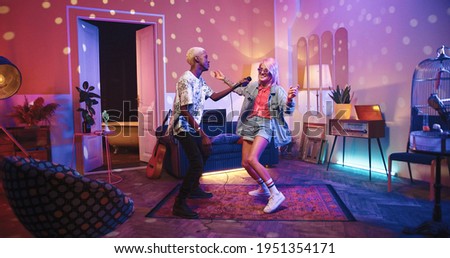 Two happy positive multi-ethnic youth dancing moving rhythmically in good mood at home party, male and female friends having fun energetically moving in room in neon disco light, retro style concept Royalty-Free Stock Photo #1951354171
