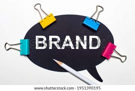 On a light background, multi-colored paper clips, a white pencil and black paper with the words BRAND. View from above