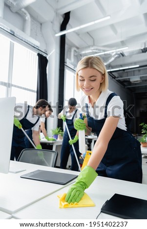 Positive worker of cleaning company cleaning table with rag near colleagues on blurred background in office