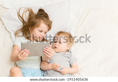 A blonde girl and sibling boy lie on bed and watch cartoons or communicate via video on smartphone. Parental control on the internet.joint leisure time. Online communication with relatives. top view