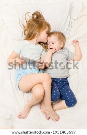 A blonde girl and sibling boy lie on bed and watch cartoons or communicate via video on smartphone.Parental control on internet.joint leisure time.Online communication with relatives.vertical.Top view