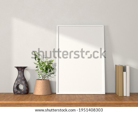 Plain white large size poster frame on the wooden table along with vase flower pot and books
