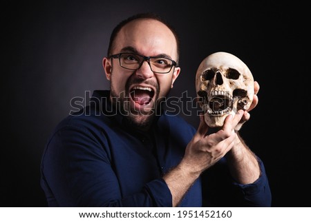 Studio portrait of a screaming man in a blue shirt with a beard and glasses, photographed against a black background and holding a human skull