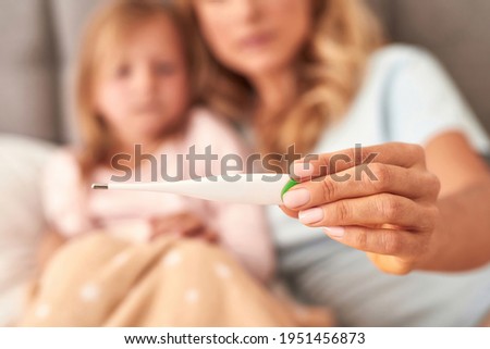 A picture of a mother checking her daughters temperature, girl is having fever. Both laying on the bed background unfocused