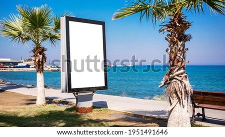 Blank lightbox with white space for mockup stands vertically on sidewalk with palm trees against blue sea and clear sky.
