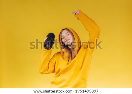 Funny young girl looking, waving hand, holding portable wireless bluetooth music speaker, isolated on yellow background. Girl in a yellow hoodie Royalty-Free Stock Photo #1951495897