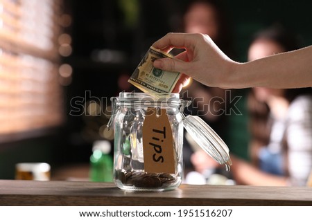 Woman putting tips into glass jar on wooden table indoors, closeup Royalty-Free Stock Photo #1951516207