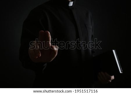 Priest with Bible making blessing gesture on dark background, closeup