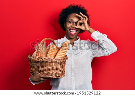 Young african american woman holding wicker basket with bread smiling happy doing ok sign with hand on eye looking through fingers 