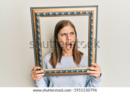 Beautiful caucasian woman holding empty frame angry and mad screaming frustrated and furious, shouting with anger. rage and aggressive concept. 