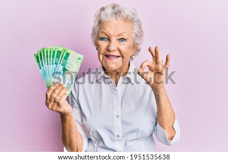 Senior grey-haired woman holding russian 200 ruble banknotes doing ok sign with fingers, smiling friendly gesturing excellent symbol 