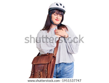 Young plus size woman wearing bike helmet and leather bag smiling with hands on chest with closed eyes and grateful gesture on face. health concept. 
