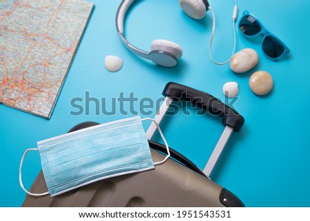 Travel during the time of COVID-19. Medical mask, suitcase, sun glasses, map, seashells on the blue background. Vacation, holidays in corona times.
