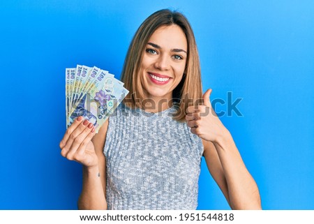 Young caucasian blonde woman holding 100 romanian leu banknotes smiling happy and positive, thumb up doing excellent and approval sign  Royalty-Free Stock Photo #1951544818