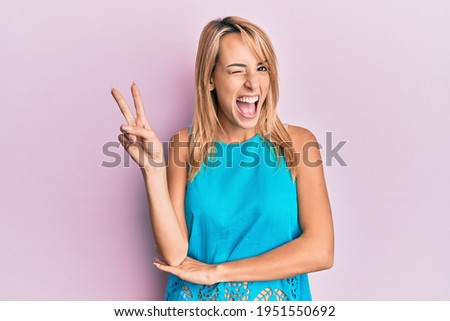 Beautiful blonde woman wearing casual clothes smiling with happy face winking at the camera doing victory sign. number two. 