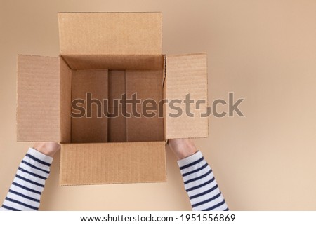 Female hands with empty open cardboard box on pastel beige color background. Top view