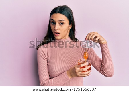 Young hispanic woman holding honey in shock face, looking skeptical and sarcastic, surprised with open mouth 
