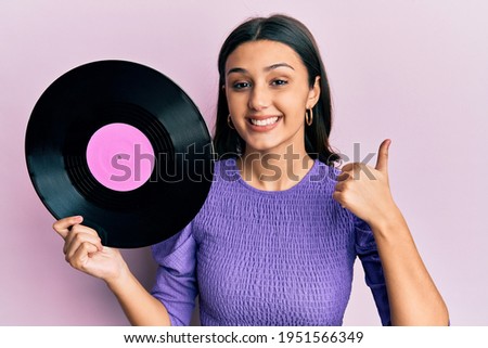 Young hispanic woman holding vinyl disc smiling happy and positive, thumb up doing excellent and approval sign 