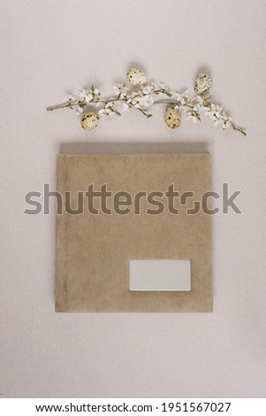 Flat lay with a beige photo album or book with a metal frame for the inscription, spring branches with white flowers and Easter eggs on a beige background. Top view, copy space
