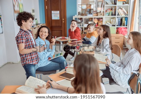 Young teacher reading with her student in front of whole class. Elementary school kids sitting on desks and reading books in classroom. Royalty-Free Stock Photo #1951567090